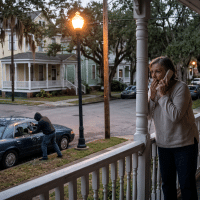 Woman on a porch speaking on a corded phone while a man works near a blue car on a residential street in a quiet neighborhood.